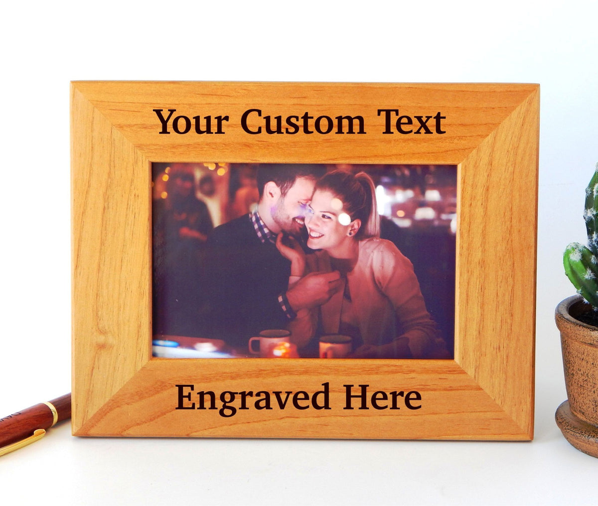 A rectangular light oak wood picture frame with engraved text placeholders reading 'Your Custom Text' at the top and 'Engraved Here' at the bottom. The frame is shown on a white surface with a small potted succulent on the right and a pen on the left.