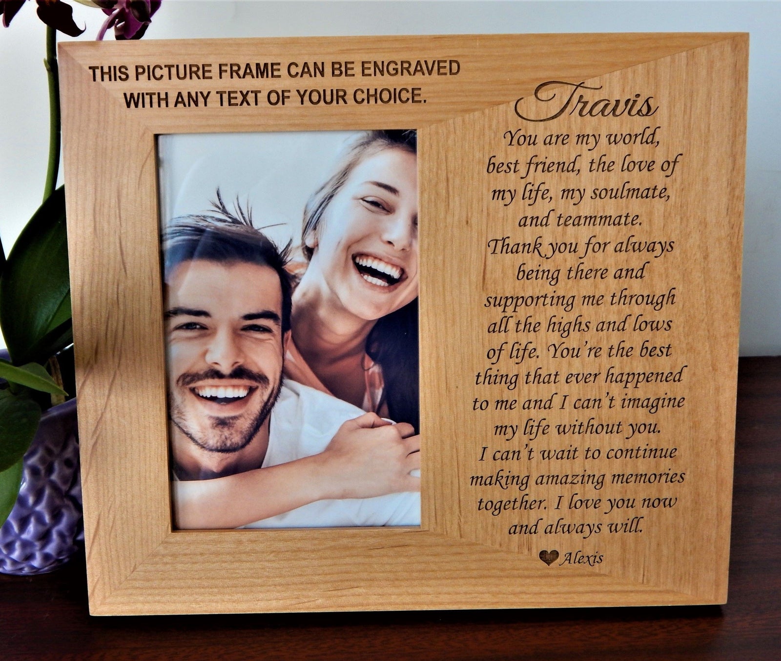 A personalized wooden picture frame engraved with customizable text, shown holding a photo of a smiling couple. The frame has a natural light oak finish and is displayed on a dark wooden surface with decorative flowers on the left side.