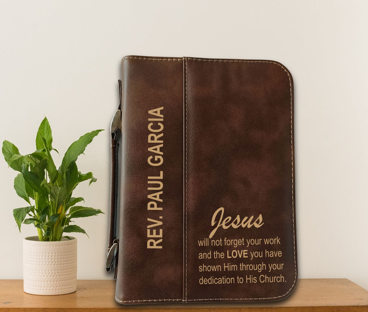A brown faux-leather Bible cover or journal with personalized gold text, standing upright on a wooden surface next to a potted green plant against a plain white wall.