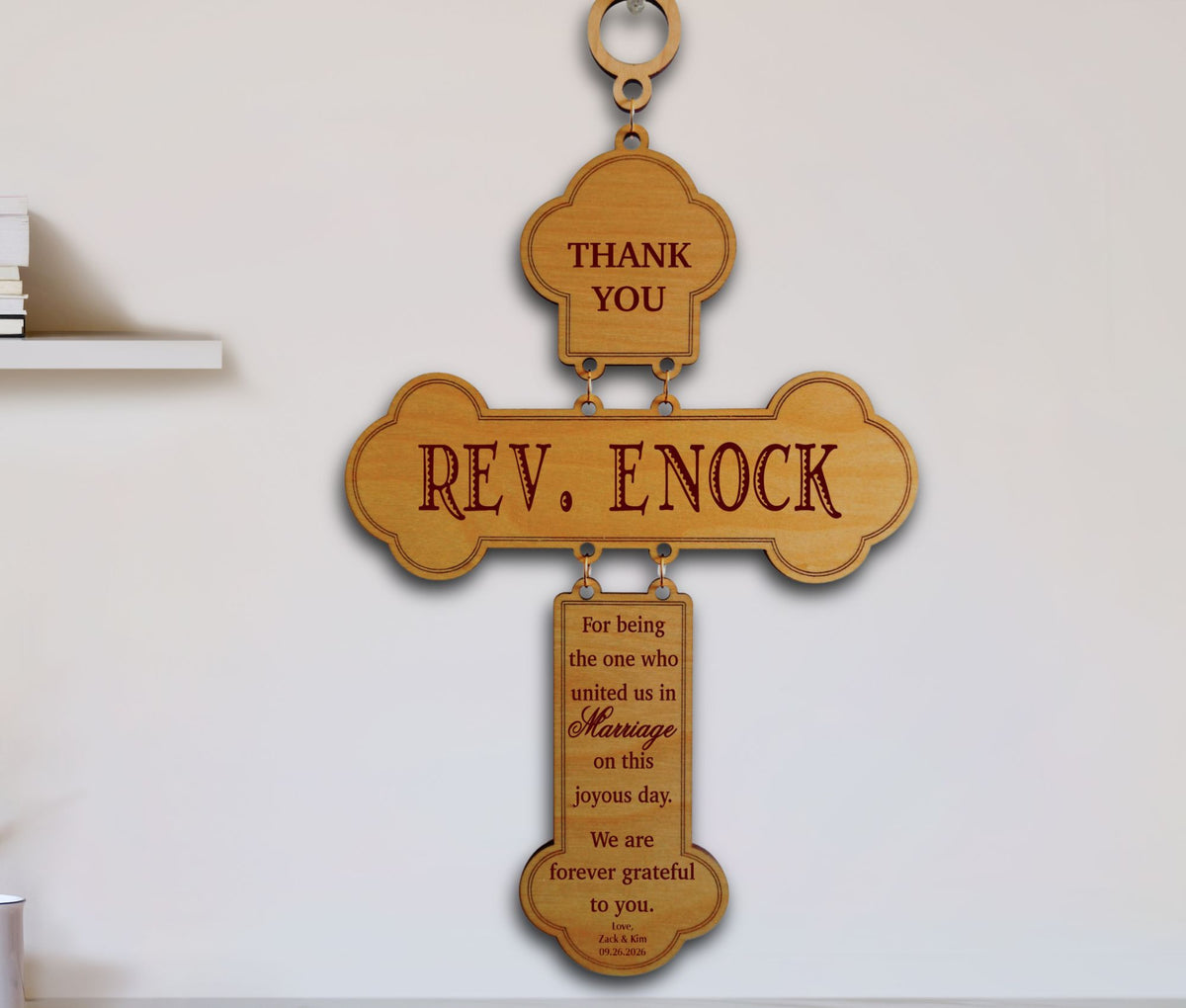 A wooden hanging cross plaque engraved with 'REV. ENOCK' and a thank you message for marriage, displayed against a plain light wall.