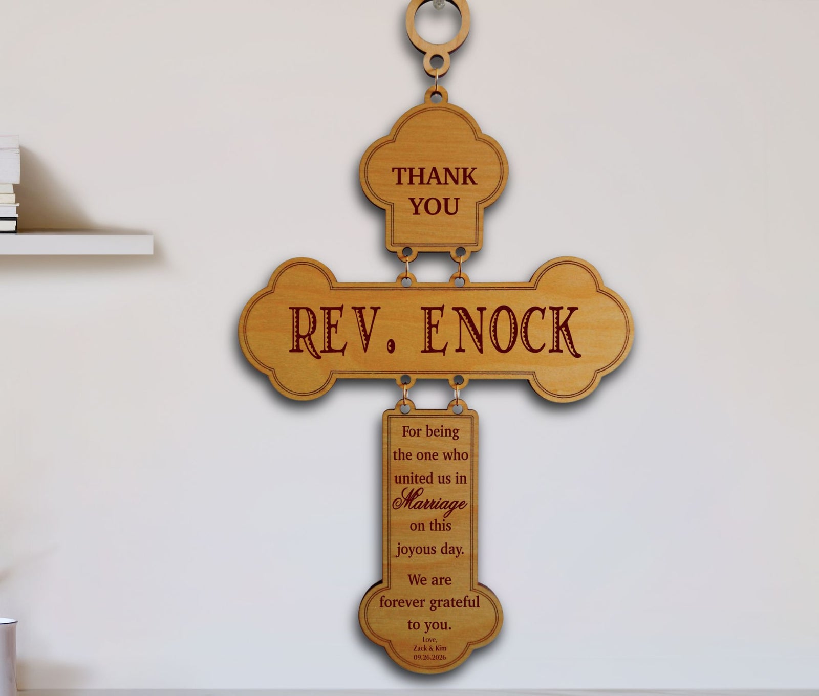 A wooden hanging cross plaque engraved with 'REV. ENOCK' and a thank you message for marriage, displayed against a plain light wall.