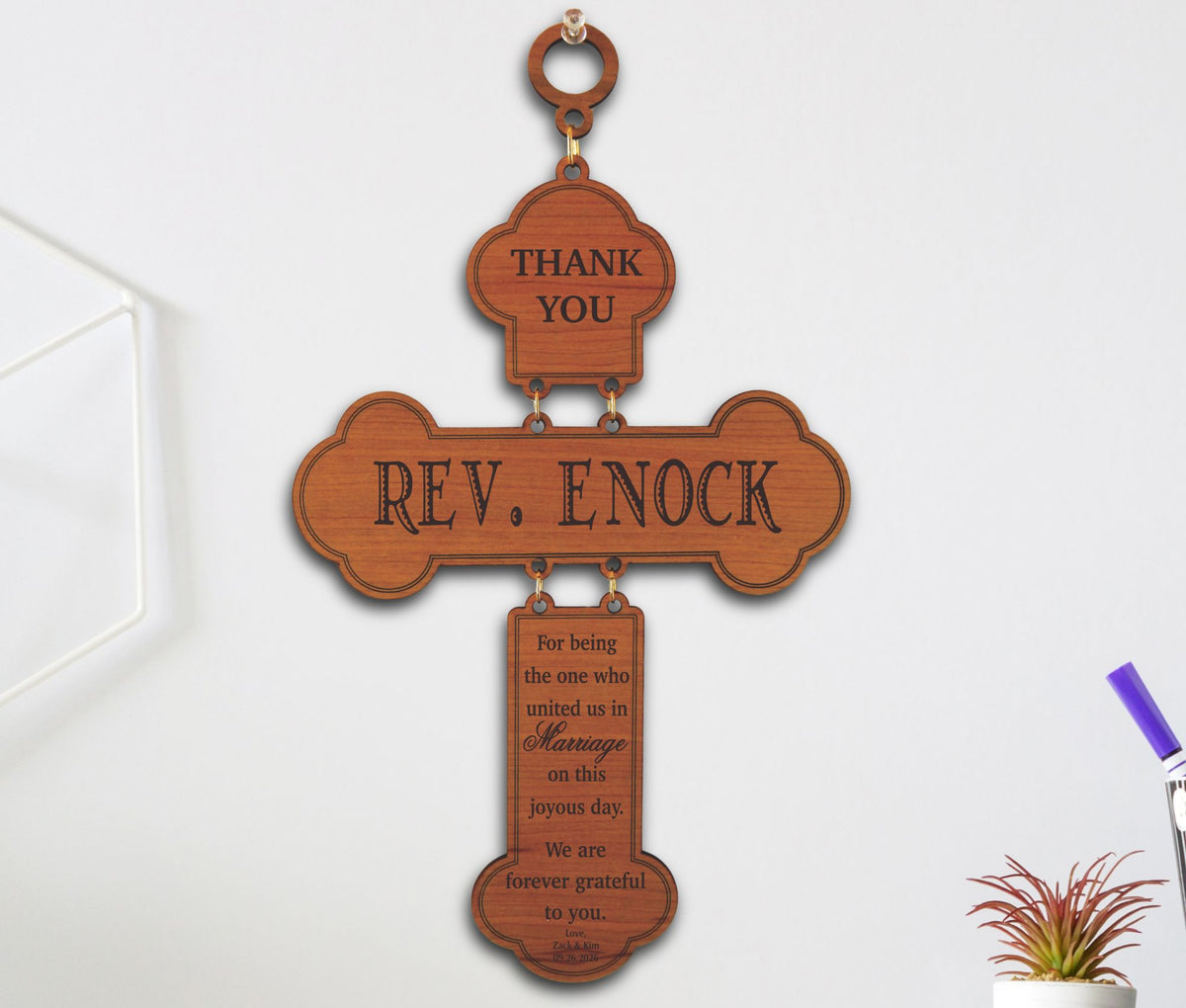 A wooden cross-shaped wall hanging with engraved wedding thank-you messages, including 'THANK YOU' and 'REV. ENOCK', shown against a light wall with small decor props at the edges.