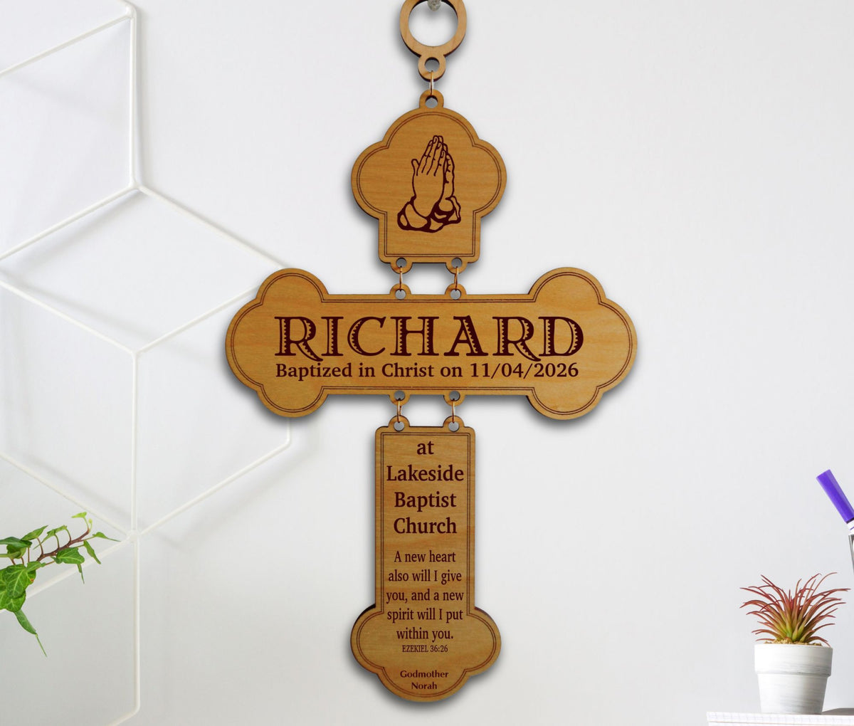 A personalized wooden baptism cross wall hanging with engraved text and a hanging loop, shown against a white wall with small plant props at the sides.