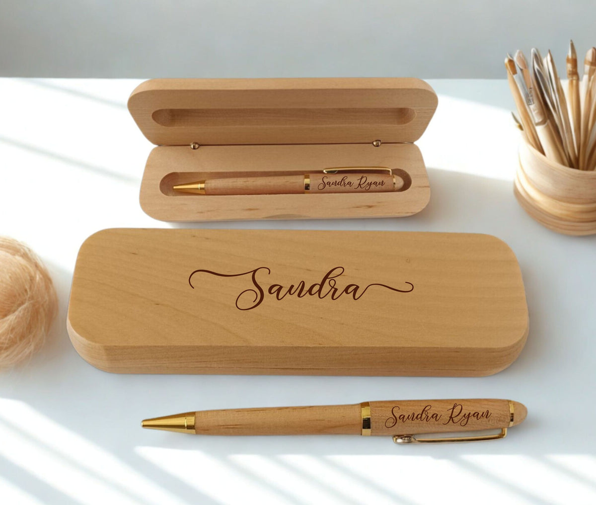 A personalized wooden pen set with a matching wooden case engraved with the name 'Sandra'. The pen and case are shown both closed and open on a white desk with soft window shadows, alongside a pencil cup in the background.