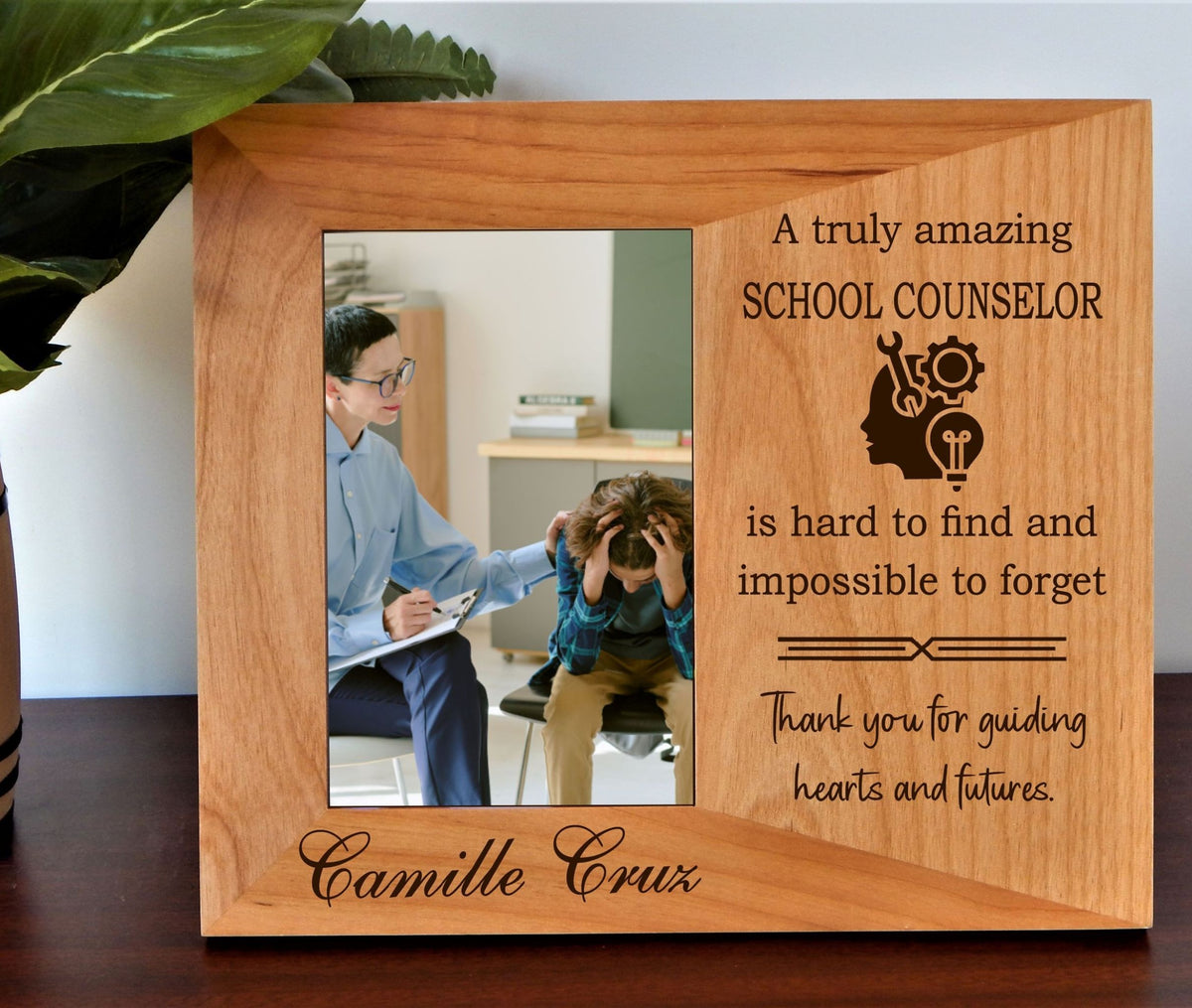 A personalized wooden photo frame with engraved text and a school counselor graphic, displayed on a dark wooden surface with a green plant leaf in the background.