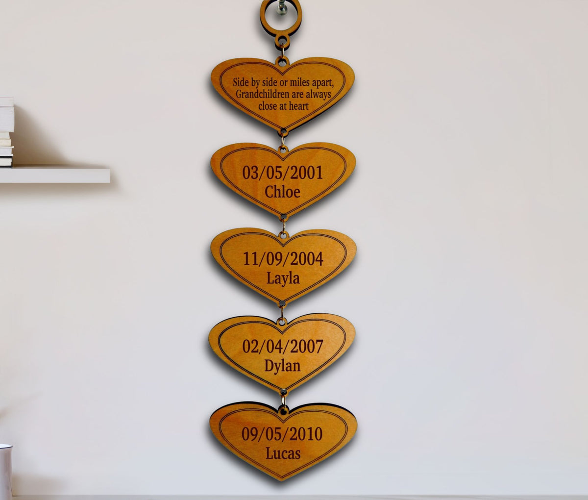 A vertical wooden hanging plaque made of five connected heart shapes, each engraved with a date and a child's name, displayed against a plain white wall.