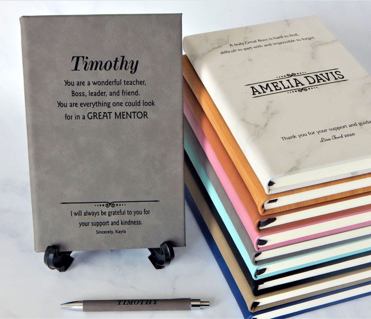 Personalized hardcover notebooks displayed on a white surface, featuring a gray notebook on small black stands, a stack of colorful notebooks on the right, and a matching gray pen in front. The notebooks are customized with names and inspirational mentorship messages.