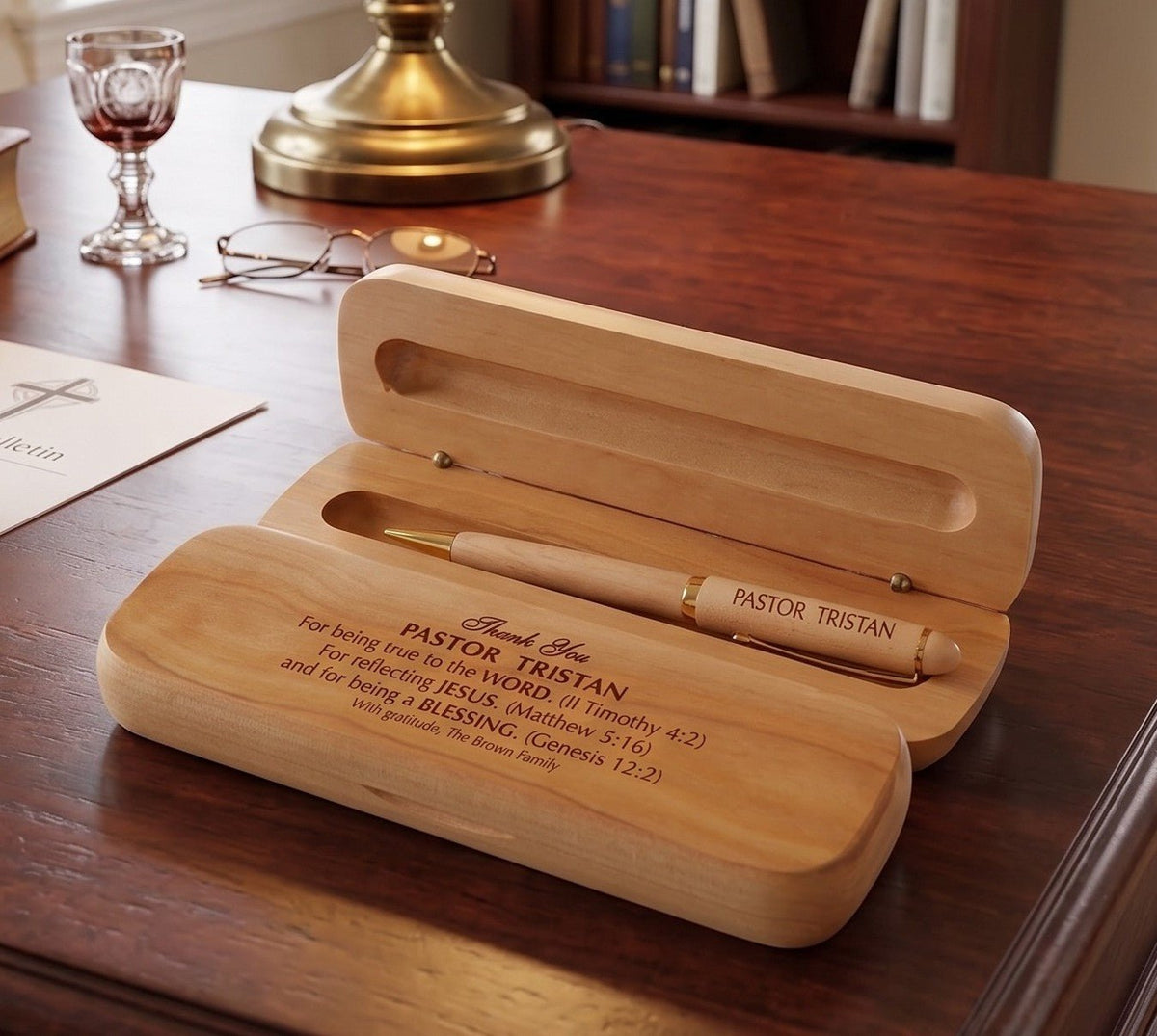 A personalized wooden pen case set on a dark wood desk, featuring an open lid holding a cream-colored pen engraved with 'PASTOR TRISTAN'. The bottom lid is engraved with a Bible verse starting with 'Thank You PASTOR TRISTAN'.