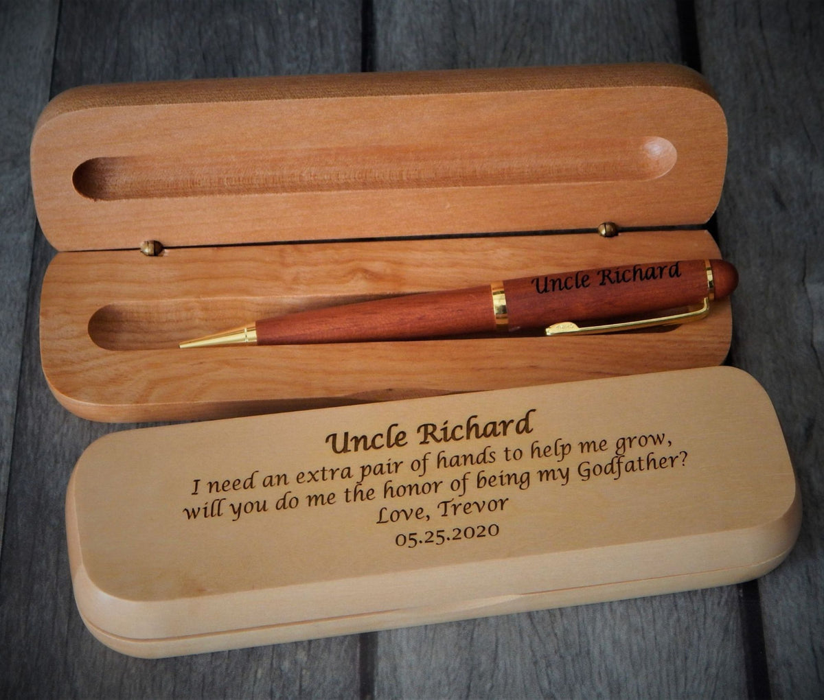 Personalized wooden keepsake box set with an engraved pen, shown on a gray wood plank background. The set includes an open light brown wooden box with a reddish-brown pen inside, and a closed matching box in front engraved with a message for 'Uncle Richard'.