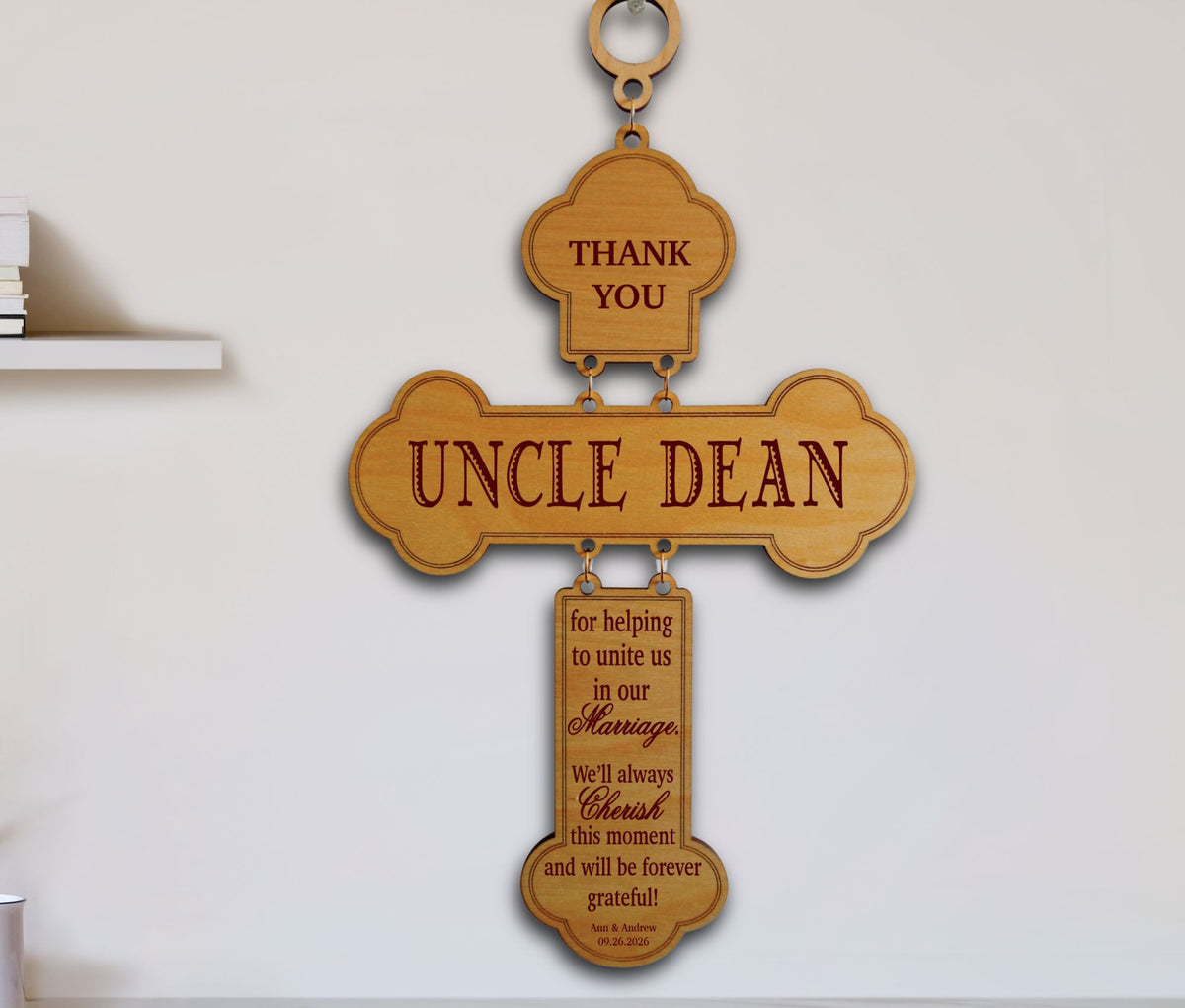A wooden hanging wall sign shaped like a cross with engraved text reading 'UNCLE DEAN' and additional memorial-style text on a smaller lower pendant, displayed against a light wall.