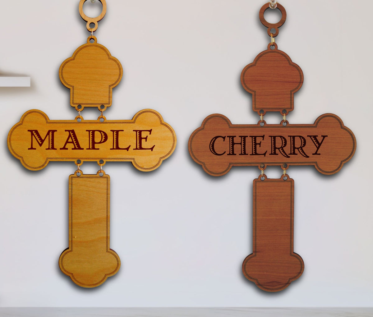 Two wooden cross wall hangings displayed side by side on a white wall. The left cross is made of light maple wood and is engraved with the word 'MAPLE'.