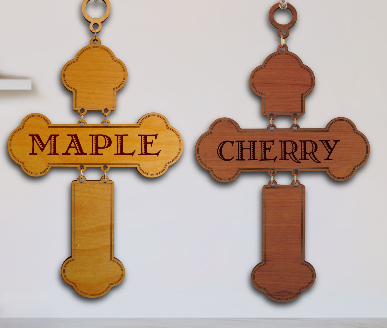 Two wooden cross wall hangings displayed side by side on a white wall. The left cross is made of light maple wood and is engraved with the word 'MAPLE'.