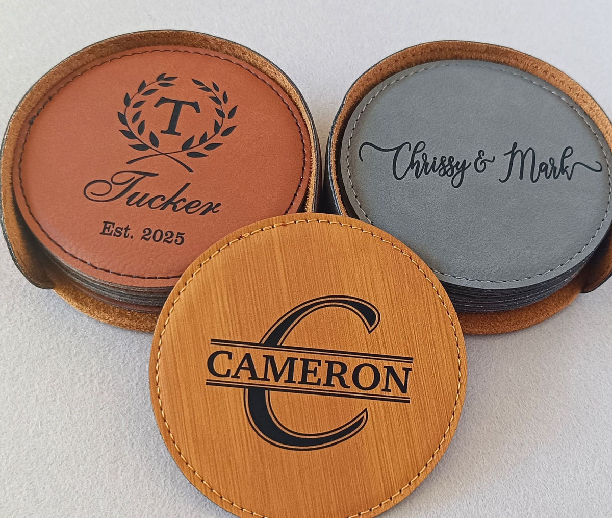 Three round personalized leather coasters displayed on a light neutral background. One coaster in the front is light tan wood with a black monogram 'C' and the name 'CAMERON'.