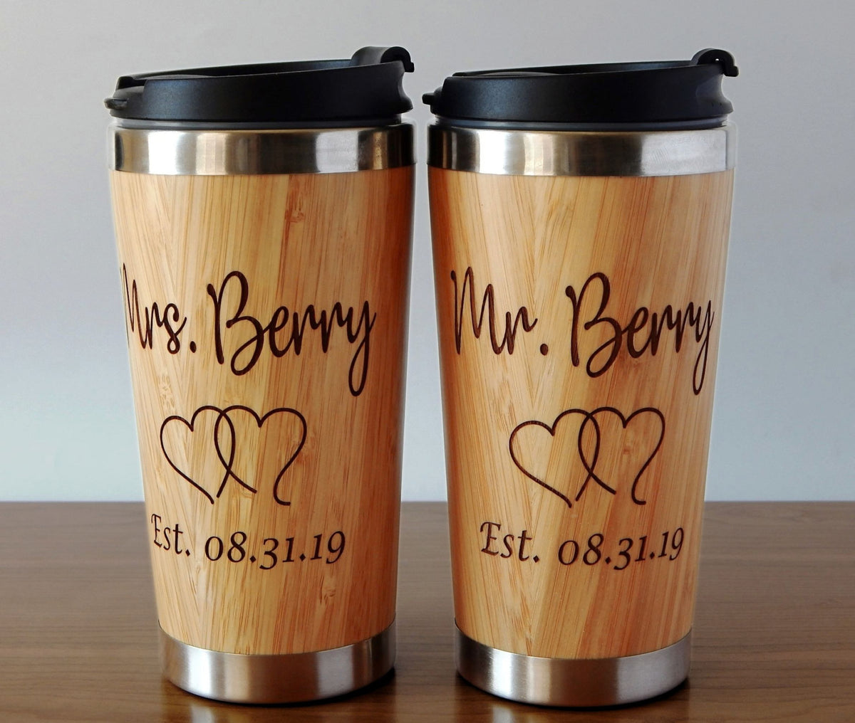 Two personalized stainless steel travel mugs with a light wood grain finish, featuring black lids and engraved names 'Mrs. Berry' and 'Mr.