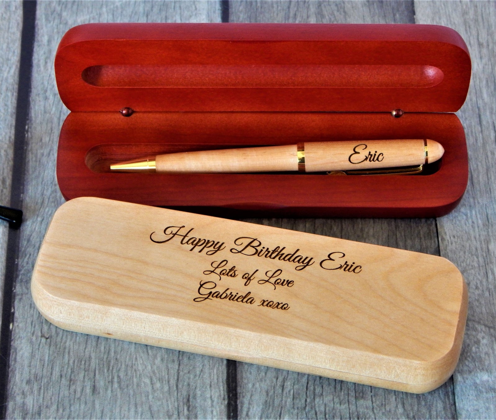 A personalized wooden pen set displayed on a gray wood background, featuring an open reddish-brown wooden presentation box with a light wood pen inside, and a matching closed light wood box in front engraved with 'Happy Birthday Eric, Lots of Love, Gabriela xxxa'.