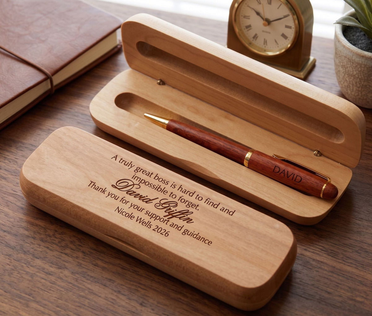 Personalized wooden pen case set on a wooden desk, featuring an open light wood case holding a reddish-brown wooden pen with gold trim, alongside a closed matching case engraved with a message. A clock, notebook, and small plant are softly blurred in the background.