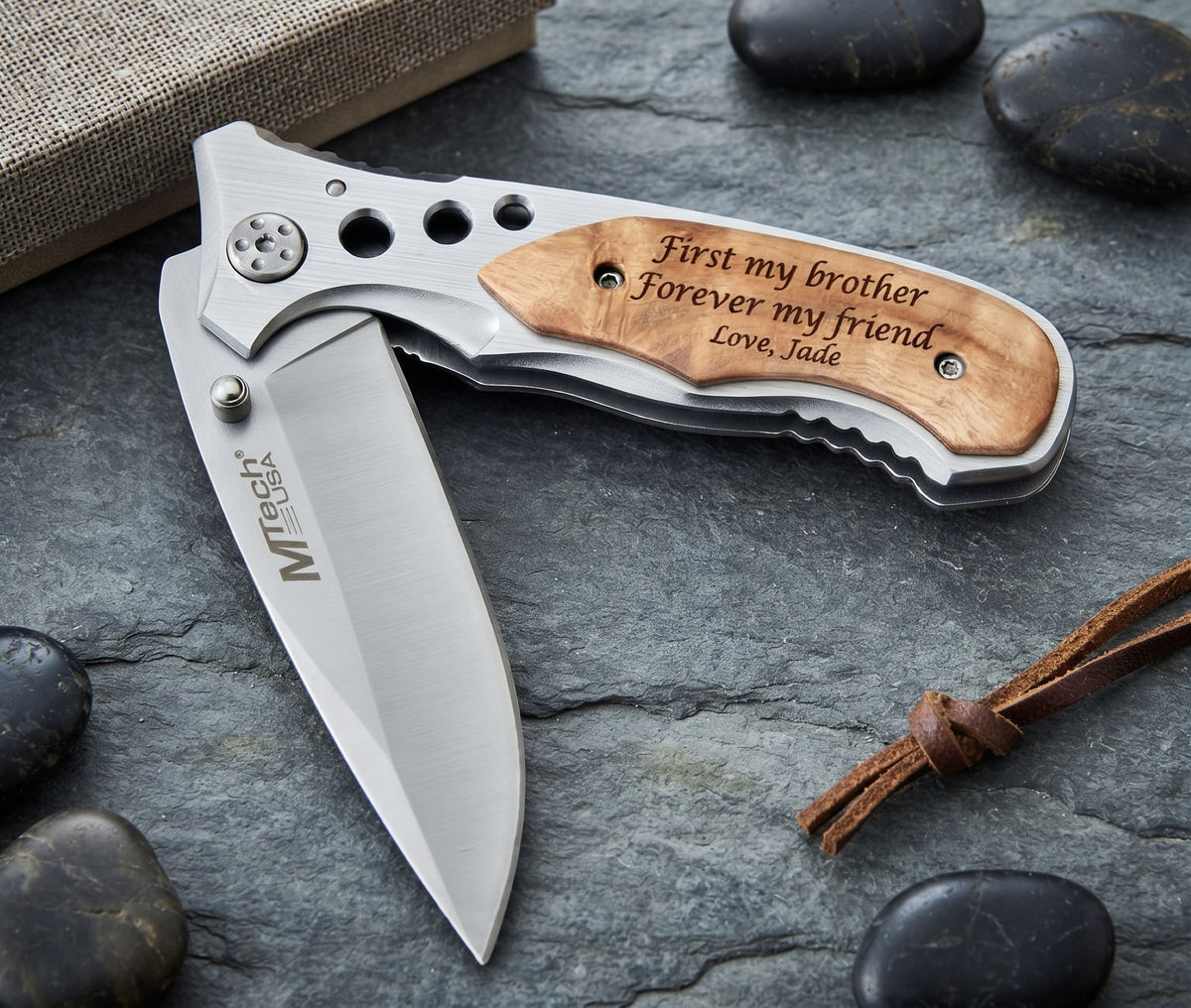 A folding pocket knife with a silver stainless steel blade and handle, featuring a natural wood inlay engraved with 'First my brother Forever my friend Love, Jade'. The knife is displayed on a dark gray slate surface with decorative stones and a leather cord.