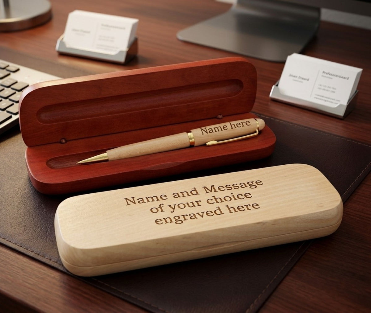 A personalized wooden pen gift set displayed on a dark desk, featuring an open reddish-brown wooden presentation box with a gold-accented pen inside, and a light natural wood engraved case in the foreground.