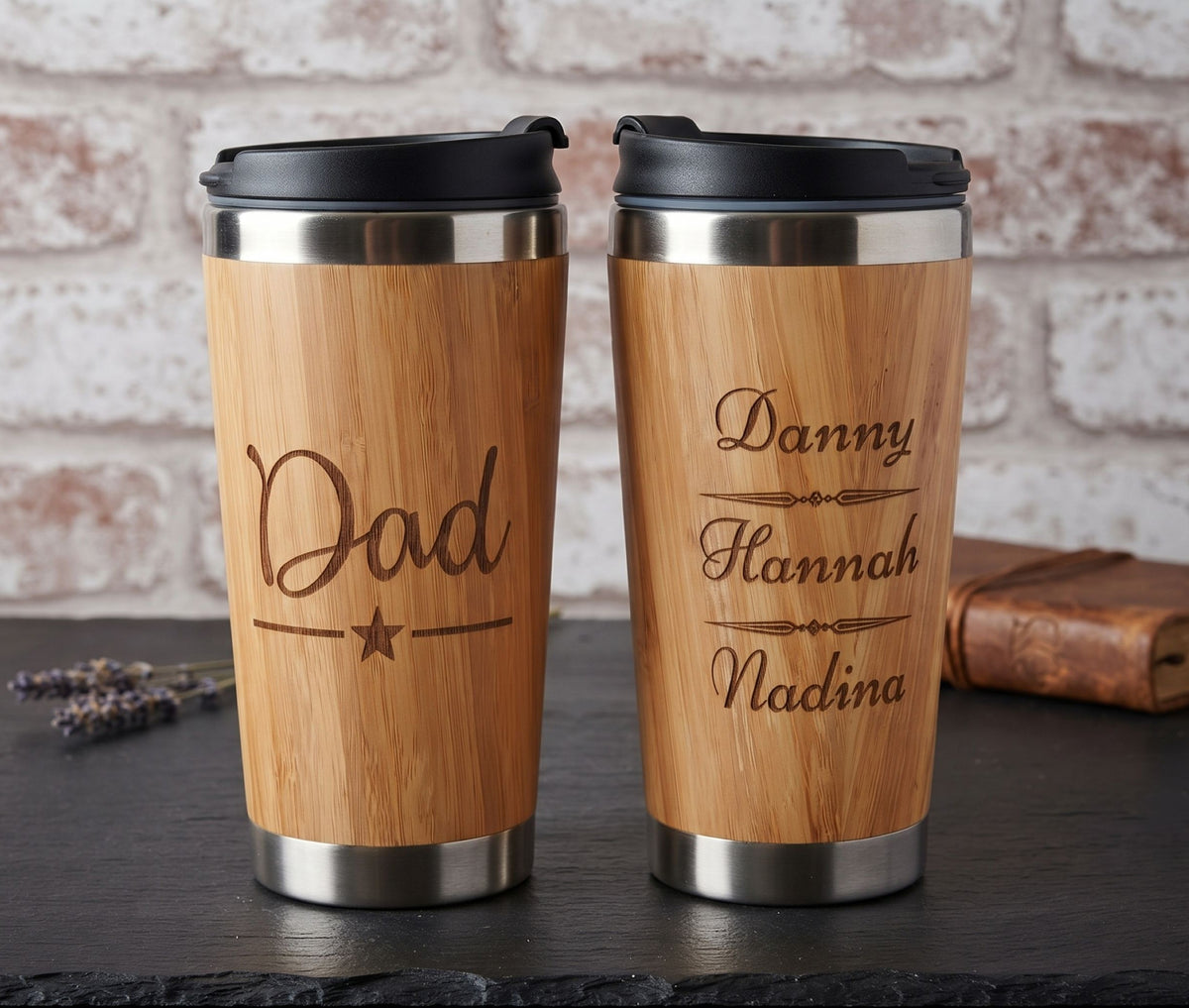 Two personalized bamboo travel mugs with black lids and stainless steel trim, displayed side-by-side on a dark tabletop against a rustic white brick wall. One mug is engraved with 'Dad' and the other with 'Danny Hannah Nadina'.