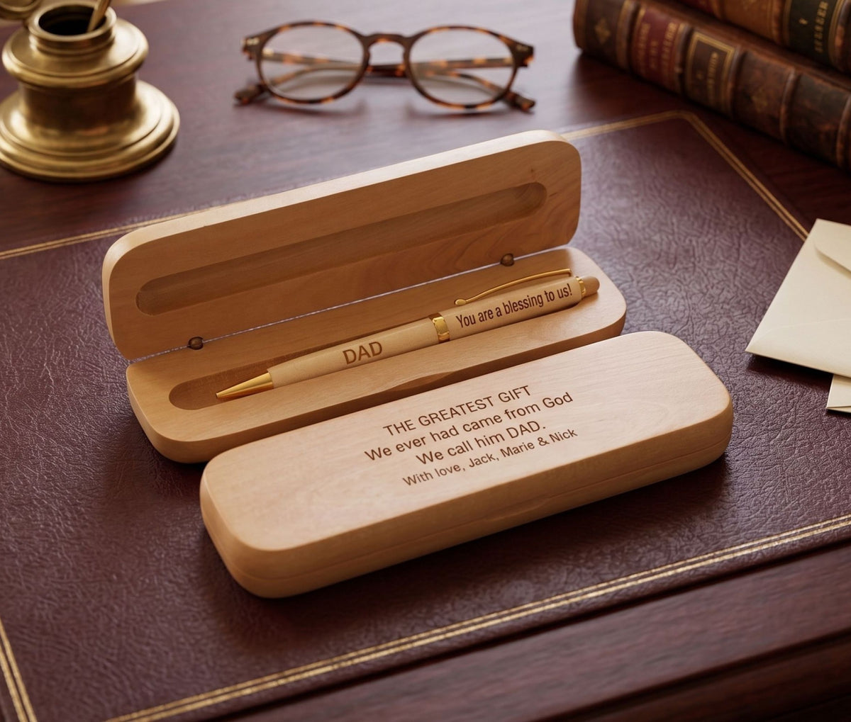 A personalized wooden pen gift set displayed on a dark brown leather desk pad, surrounded by vintage books, glasses, and a brass inkwell. The set includes an open wooden case with a light wood pen engraved with 'DAD' and 'You are a blessing to us!', and a closed wooden case engraved with 'THE GREATEST GIFT We ever had came from God We call him DAD.