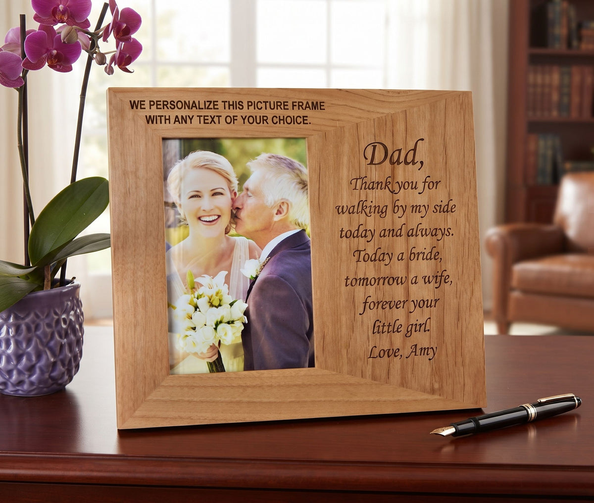 A personalized wooden picture frame with engraved text, displayed on a wooden desk next to a purple orchid and a black pen. The photo insert shows a bride and groom.