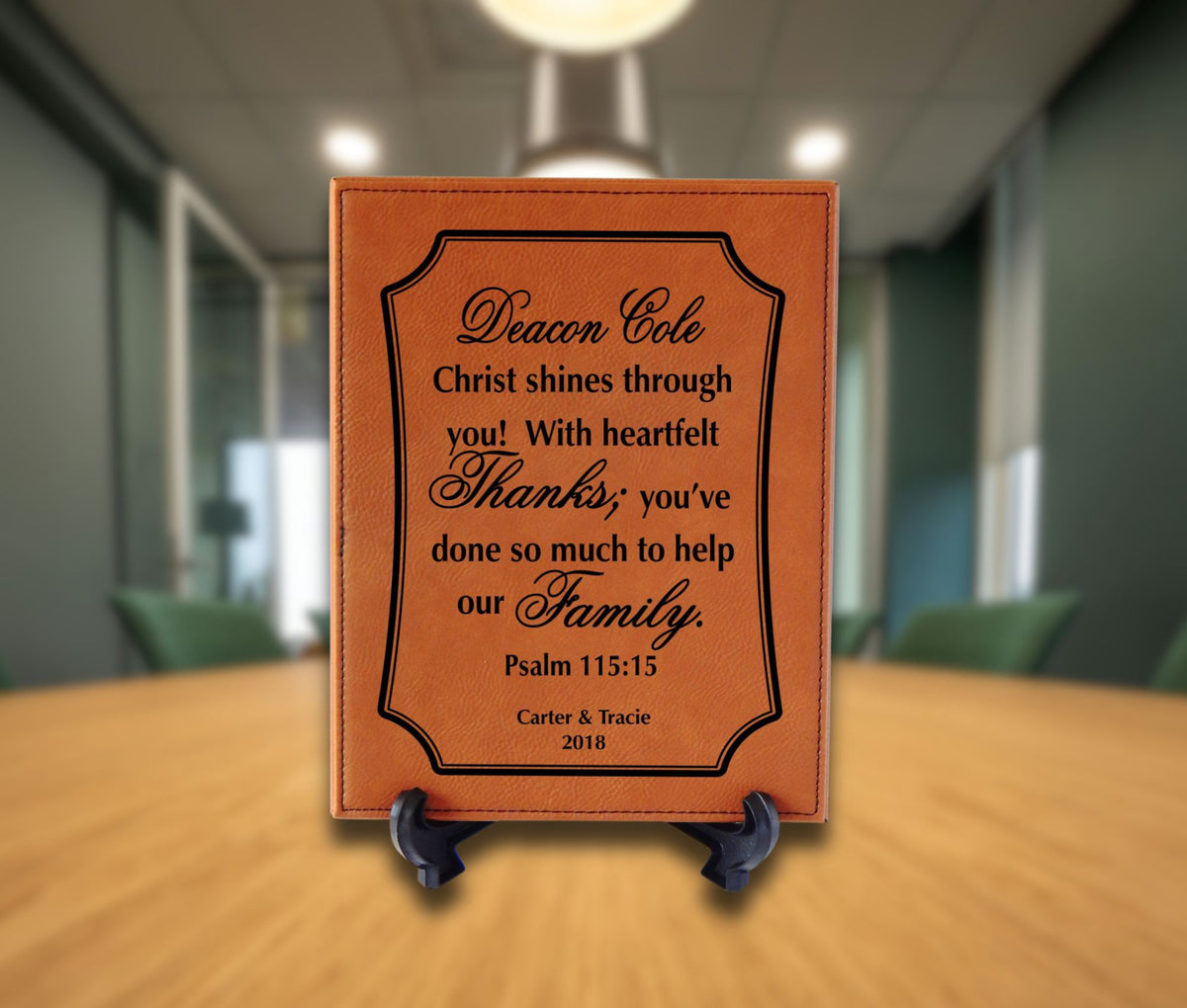 A personalized brown leather plaque with black engraved text, displayed on a small black easel stand on a wooden table in a blurred office setting.
