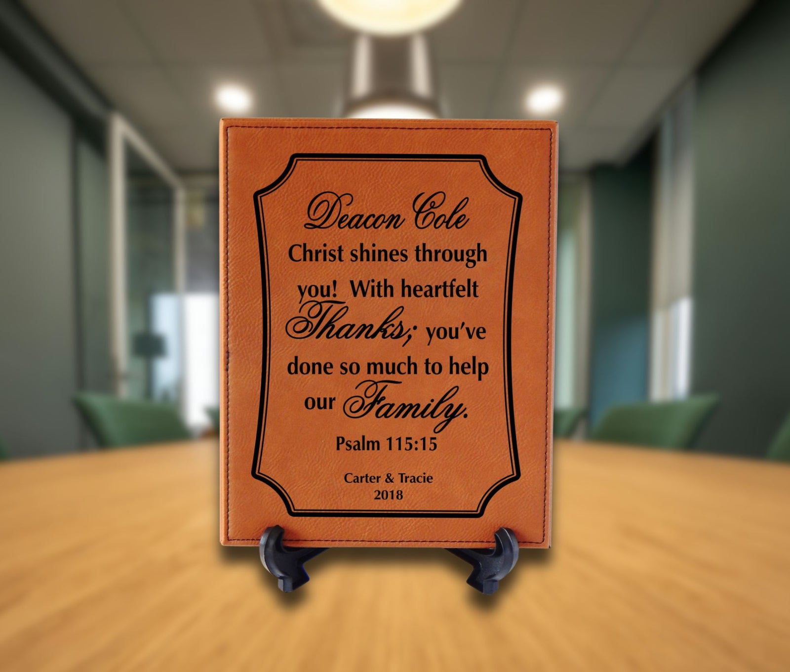 A personalized brown leather plaque with black engraved text, displayed on a small black easel stand on a wooden table in a blurred office setting.