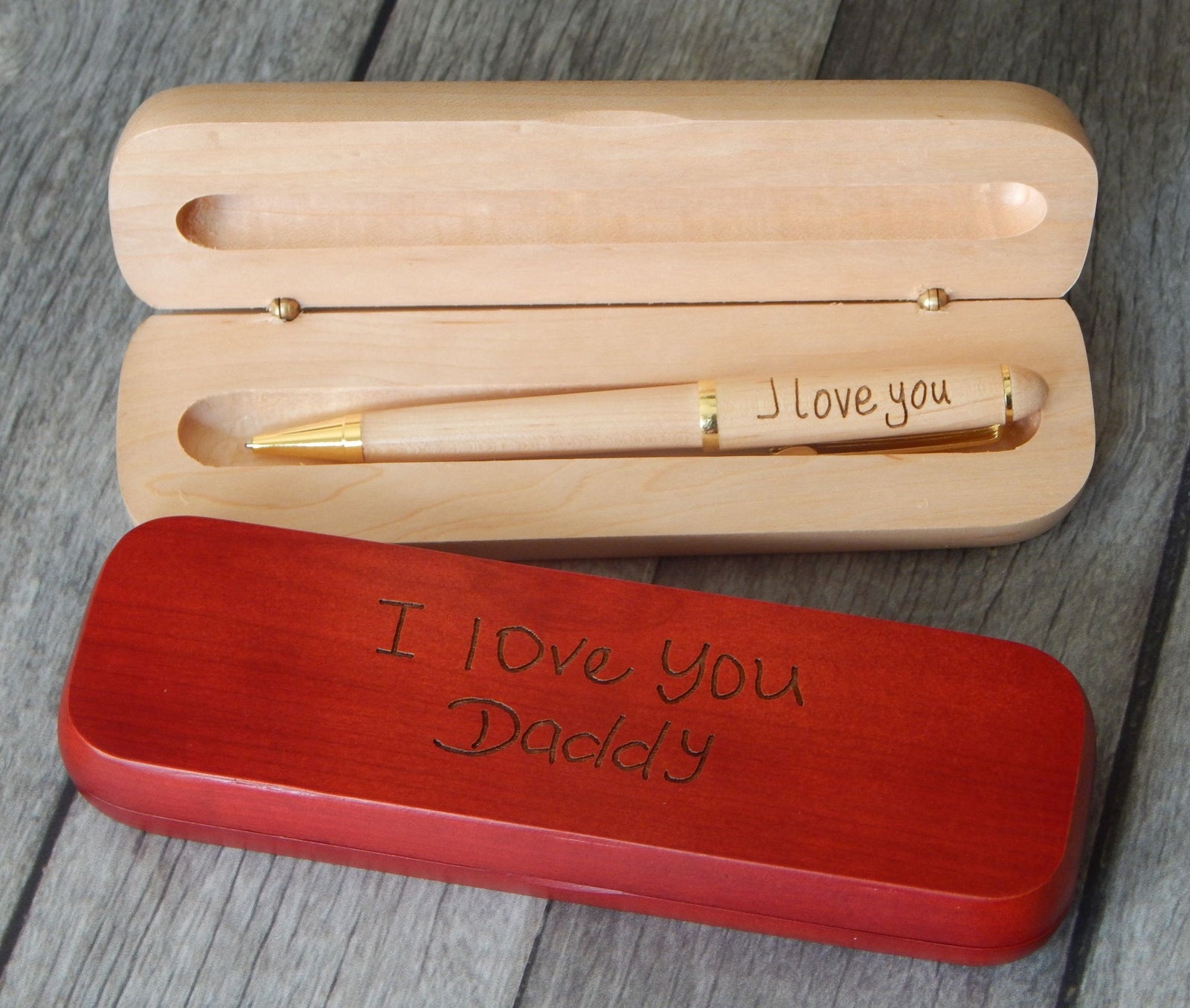 A personalized wooden pen gift set on a gray wood background, featuring an open natural wood box with a wooden pen engraved with 'I love you' and a red wooden case engraved with 'I love you Daddy'.