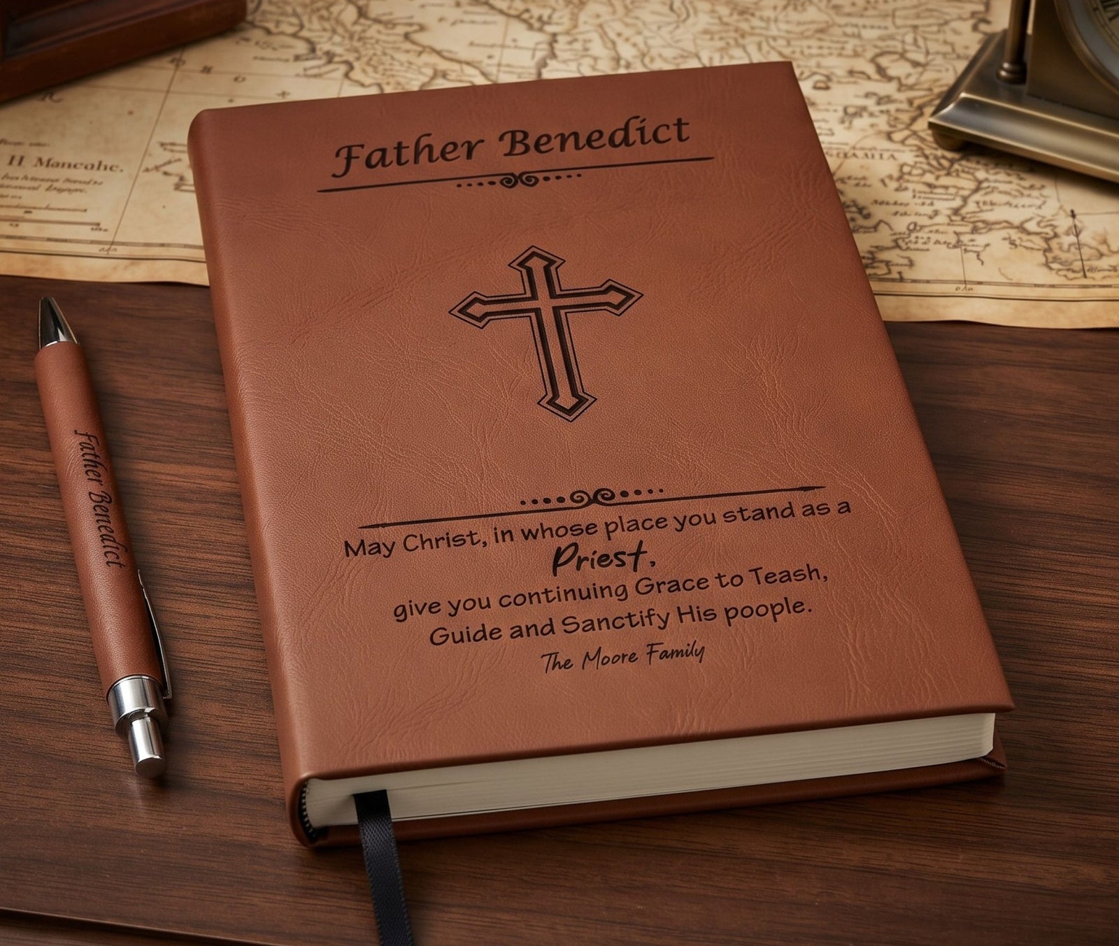 A personalized brown faux-leather Bible or prayer journal lies open on a wooden desk, showing the cover engraved with the name 'Father Benedict' and a cross. A matching brown pen with the same personalization rests beside it, with an antique-style map in the background.
