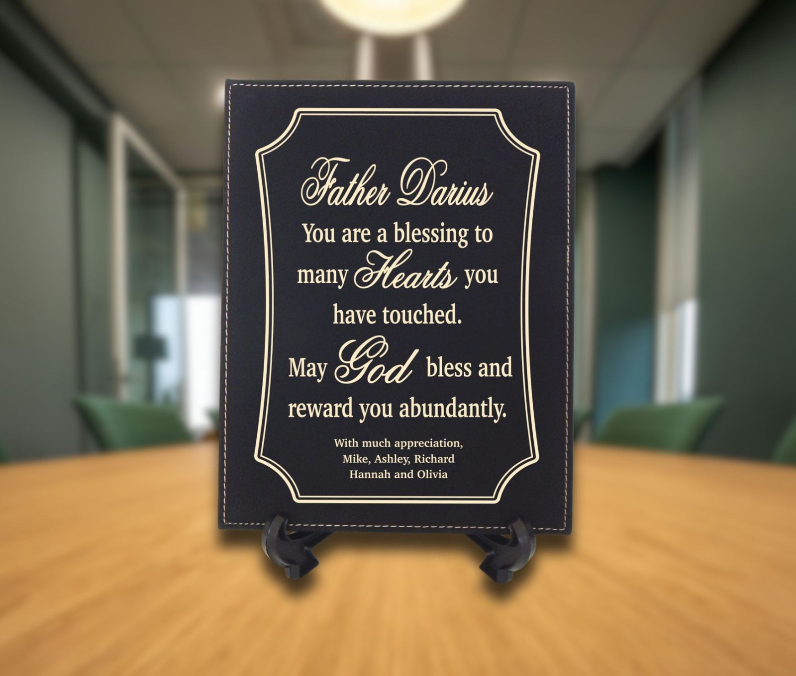A personalized black memorial plaque with gold lettering, displayed on a small black easel stand on a wooden table in a blurred office hallway setting.