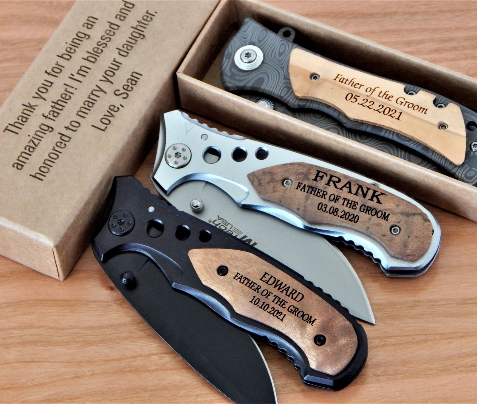 Three personalized folding pocket knives displayed on a wooden surface with their kraft gift boxes. The knives feature black, silver, and wood-grain handles with laser-engraved names and dates, including 'FRANK', 'EDWARD', and 'PATRICK - FATHER OF THE GROOM'.
