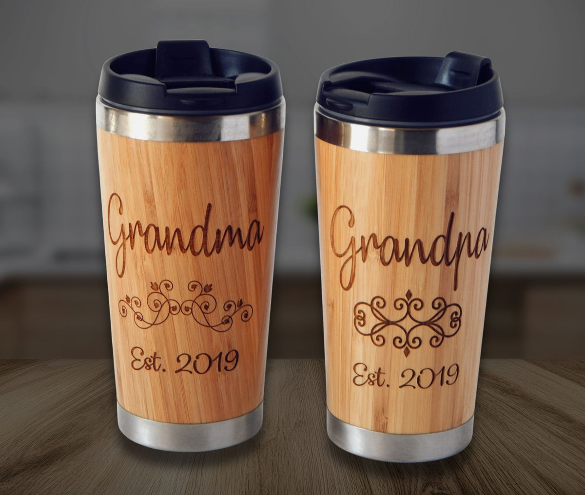 Two personalized bamboo travel tumblers with dark blue lids, standing side by side on a wooden table. Each tumbler features a laser-engraved design with the words 'Grandma' and 'Grandpa' along with 'Est.
