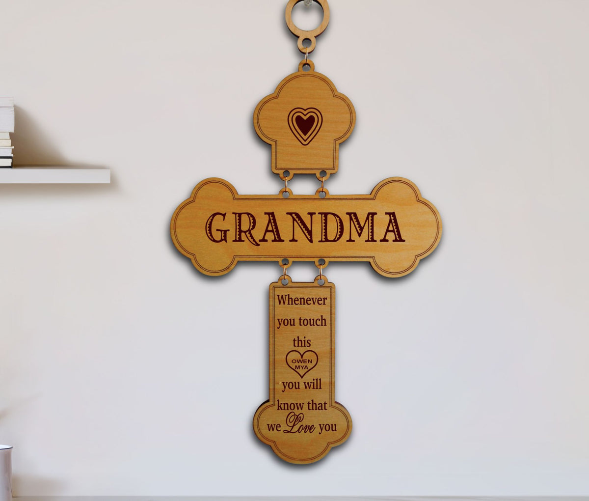 A wooden cross-shaped wall hanging engraved with the word GRANDMA and a hanging tag that reads, 'Whenever you touch this you will know that we Love you,' shown against a plain light wall.