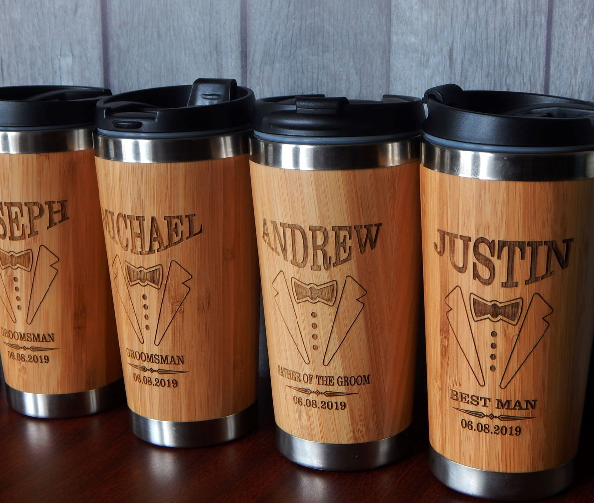 Four personalized bamboo travel mugs with black lids lined up on a dark wooden surface against a gray wood-paneled background. Each mug features an engraved tuxedo graphic and the name of a groom's friend along with wedding details.