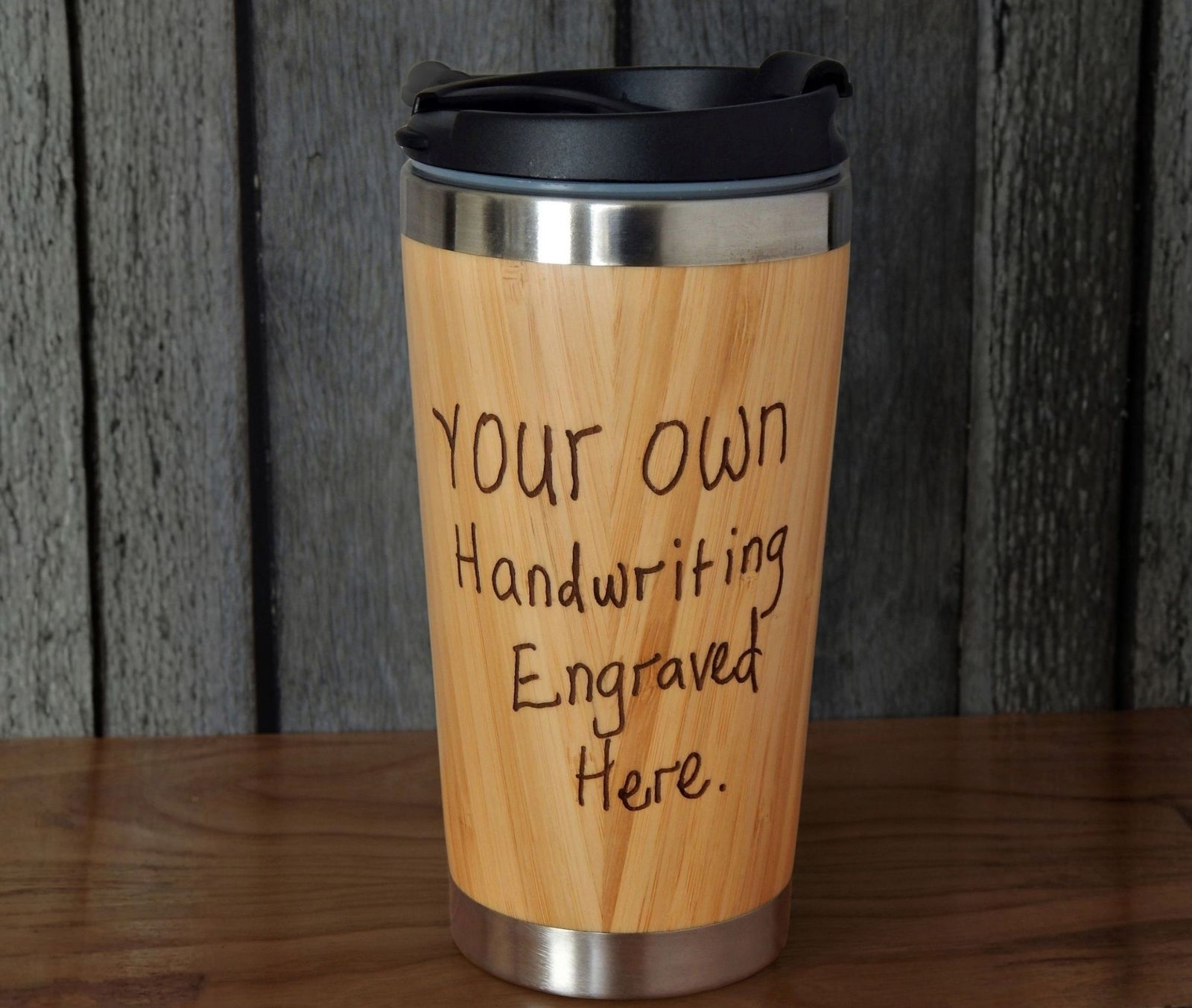 A bamboo travel tumbler with a black lid, featuring the text 'YOUR OWN Handwriting Engraved Here.' in black lettering, set against a dark wood paneled background.