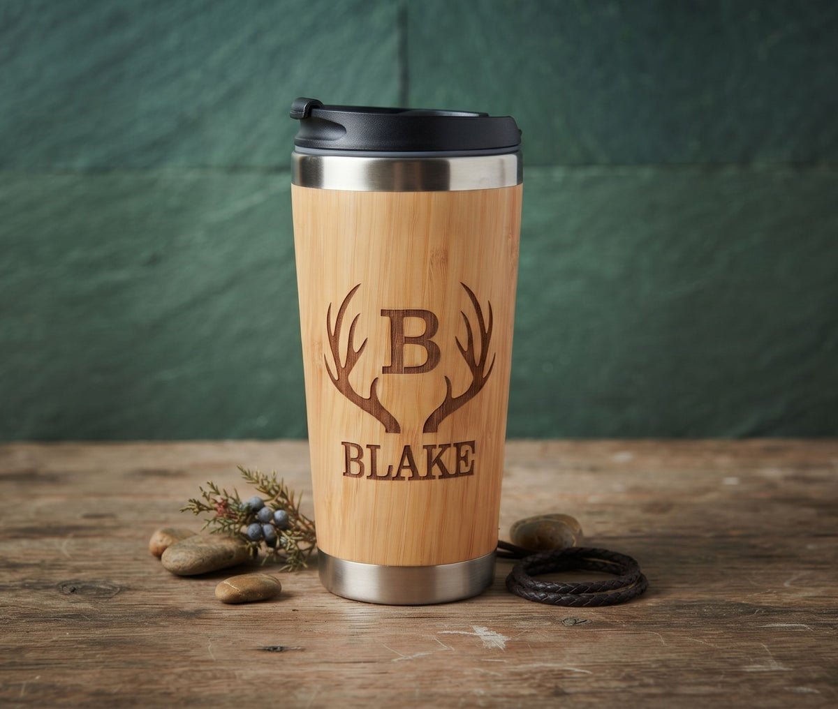 A personalized bamboo travel tumbler with a black lid, featuring a laser-engraved antler monogram 'B' and the name 'BLAKE'. The tumbler is displayed on a rustic wooden table with small decorative stones and a leather cord, set against a dark green textured background.