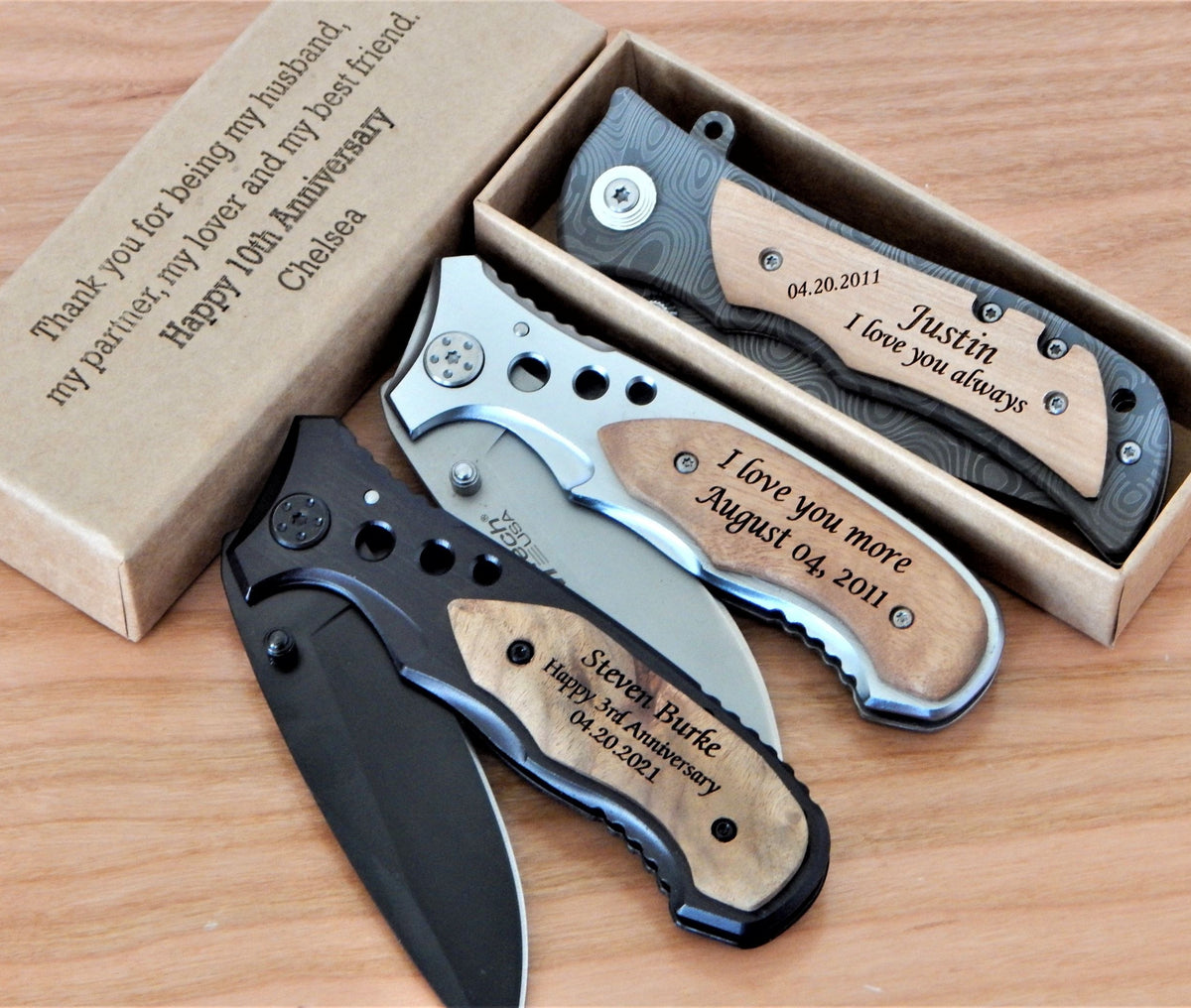 Three personalized folding pocket knives with wood-grain handles are displayed on a wooden surface next to a brown cardboard gift box with engraved anniversary text. The knives feature black, silver, and wood-grain (brown) handle colors.
