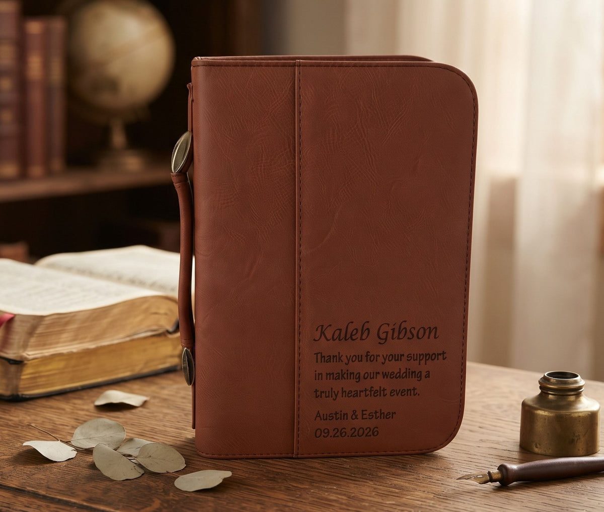 A brown leather-bound journal standing upright on a wooden table, with engraved wedding text reading 'Kaleb Gibson' and a thank-you message. The scene includes an open book, scattered leaves, and a small brass bottle in a softly lit indoor setting.