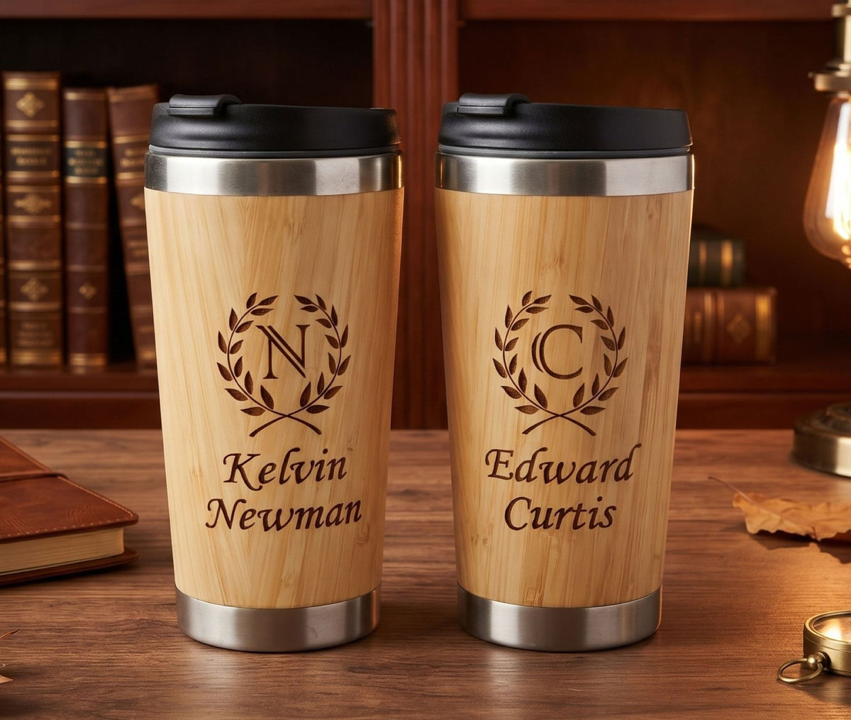 Two personalized bamboo travel tumblers with black lids and stainless steel accents, standing on a wooden table in front of a bookshelf. One is engraved with a laurel wreath and the name Kelvin Newman, and the other with a laurel wreath and the name Edward Curtis.