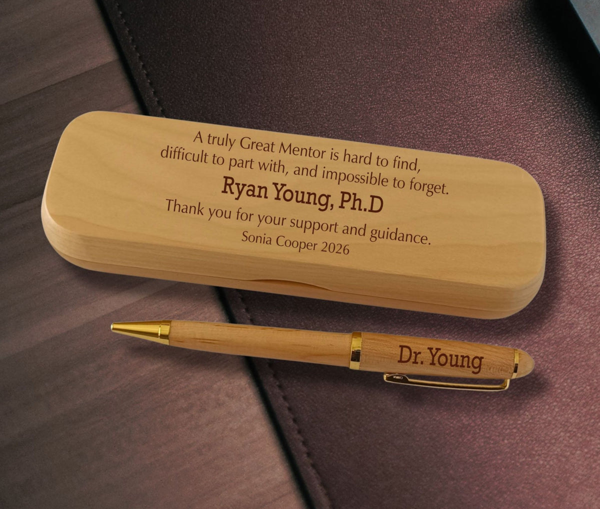 A personalized wooden pen and matching wooden case with engraved text, displayed on a dark brown leather surface. The pen is engraved with 'Dr.