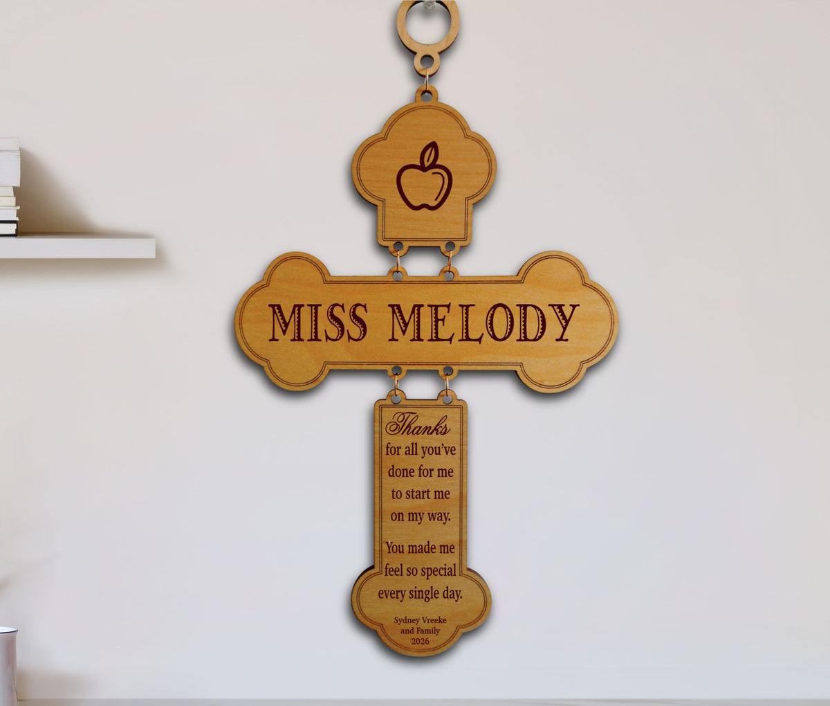 A wooden cross-shaped wall hanging with engraved text reading 'MISS MELODY' and a smaller plaque below with a motivational message. The top section shows an apple icon.