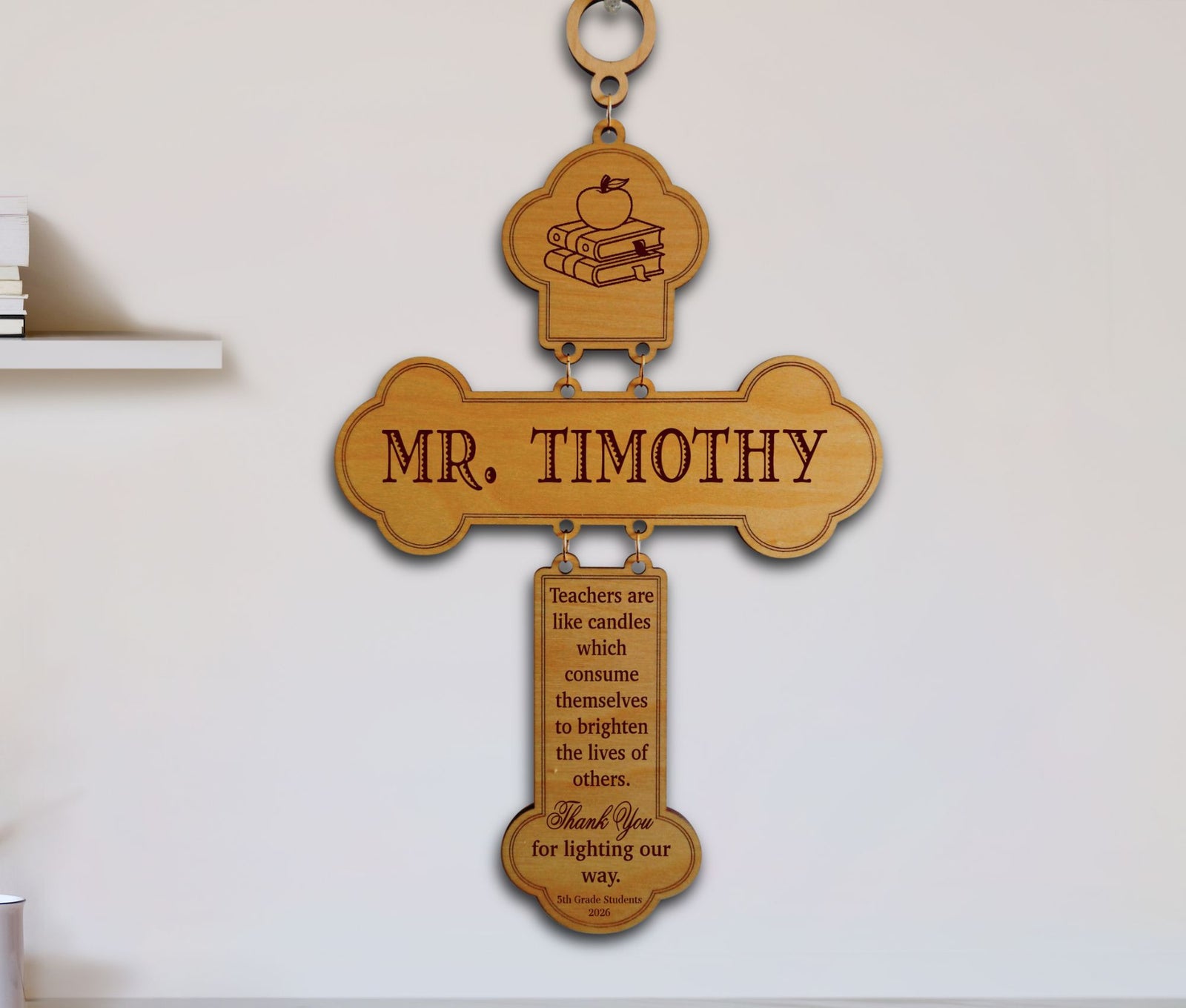 A personalized wooden cross wall hanging with the name 'MR. TIMOTHY' engraved in the center, a small book-and-apple emblem at the top, and a hanging plaque with a quote below, shown mounted on a white wall.