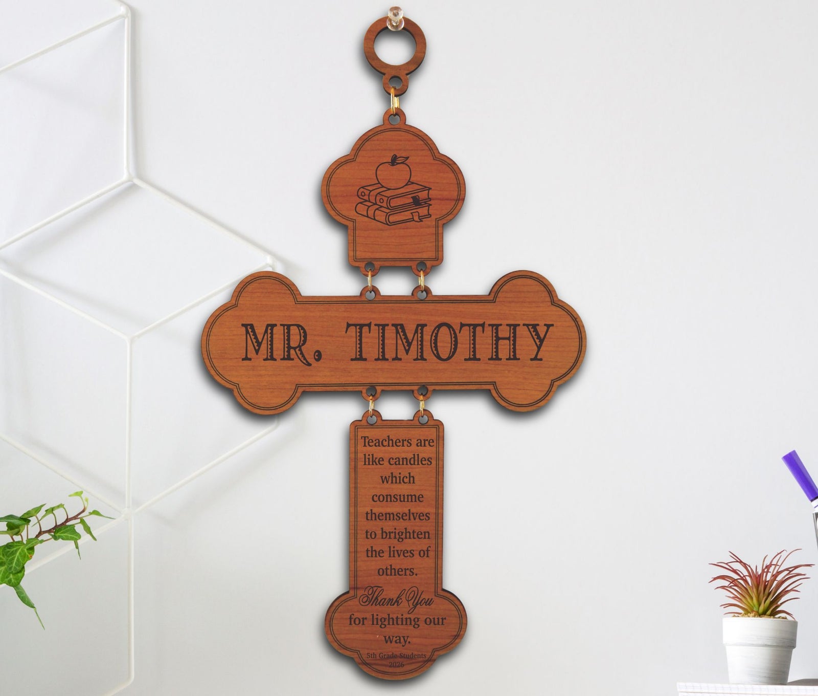 A personalized wooden wall plaque hanging on a white wall. The cross-shaped sign features a top decorative tab with a small book-and-apple illustration, a central label reading 'MR.