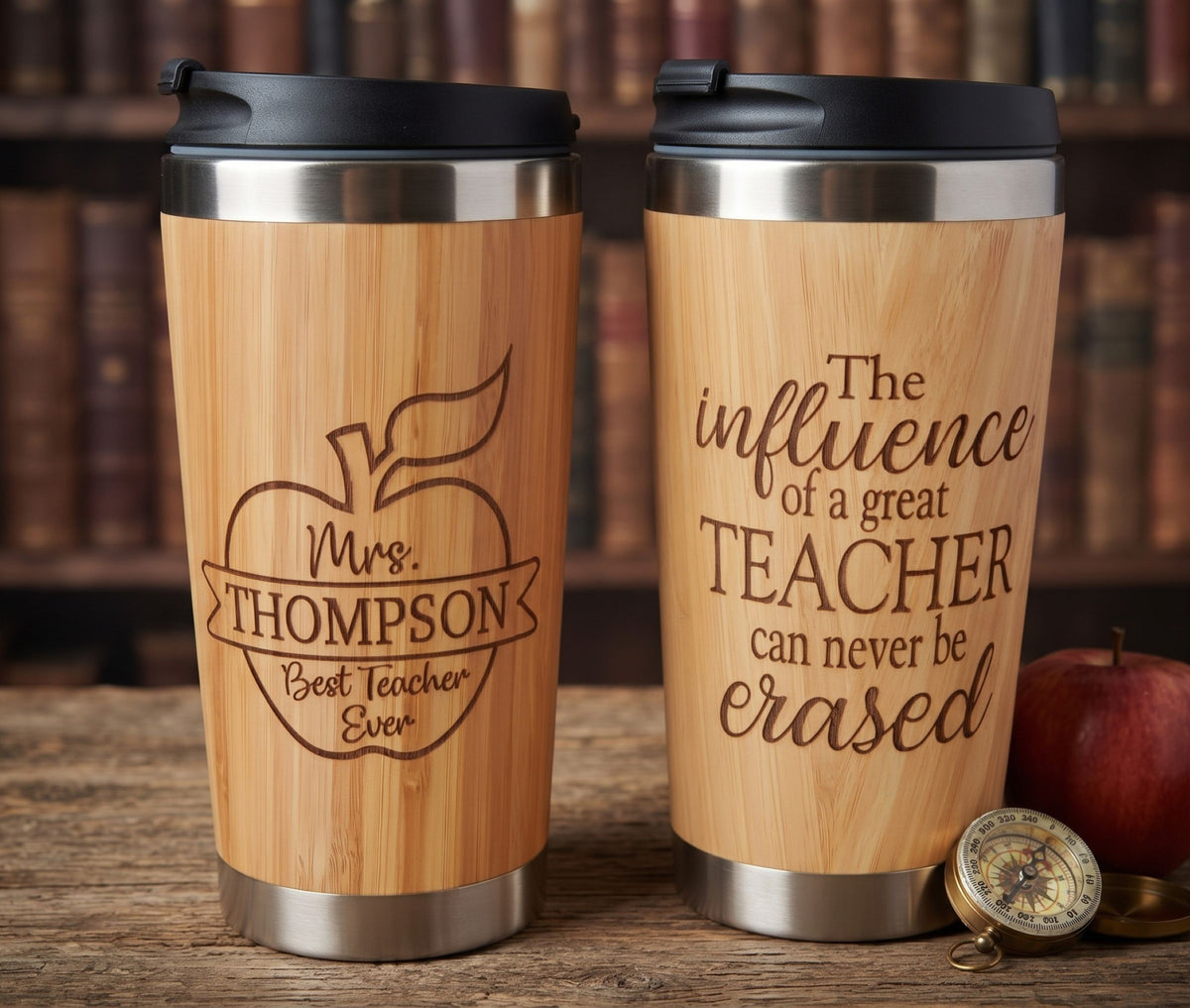 Two personalized bamboo travel tumblers with black lids and stainless steel bases, displayed on a wooden table with a blurred bookshelf background. One tumbler is engraved with an apple and the text 'Mrs.