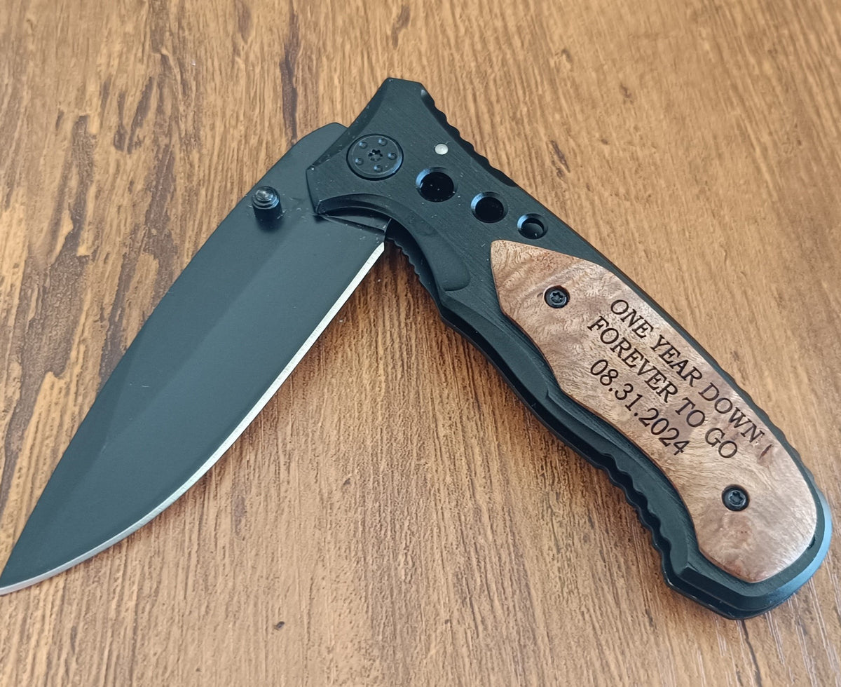 A custom pocket knife with a matte black blade and handle, featuring a natural wood inlay engraved with 'ONE YEAR DOWN FOREVER TO GO 08.31.
