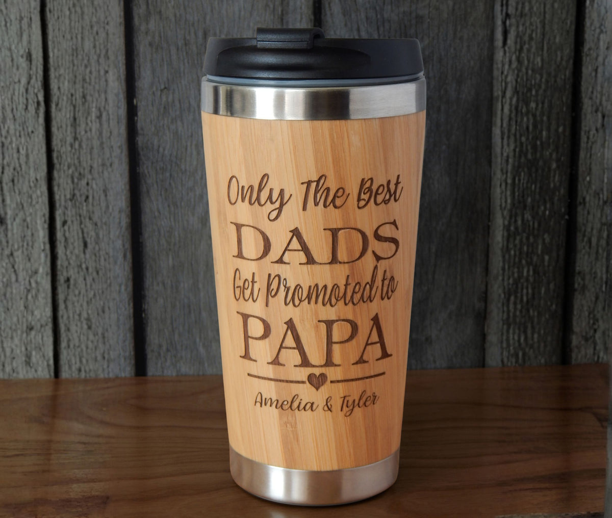 A bamboo travel tumbler with a black lid, featuring the engraved text 'Only The Best Dads Get Promoted to PAPA' and personalized names 'Amelia &amp; Tyler' on a wooden surface against a dark wood plank background.