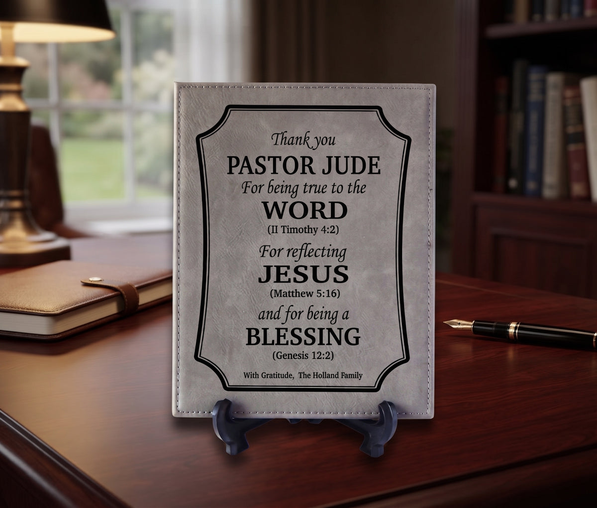 A personalized light gray leather Bible cover standing on a small black easel, featuring black printed text with Pastor Jude blessings. The book is displayed on a dark wooden desk in a warm, softly lit study with a lamp, notebook, and bookshelves in the background.