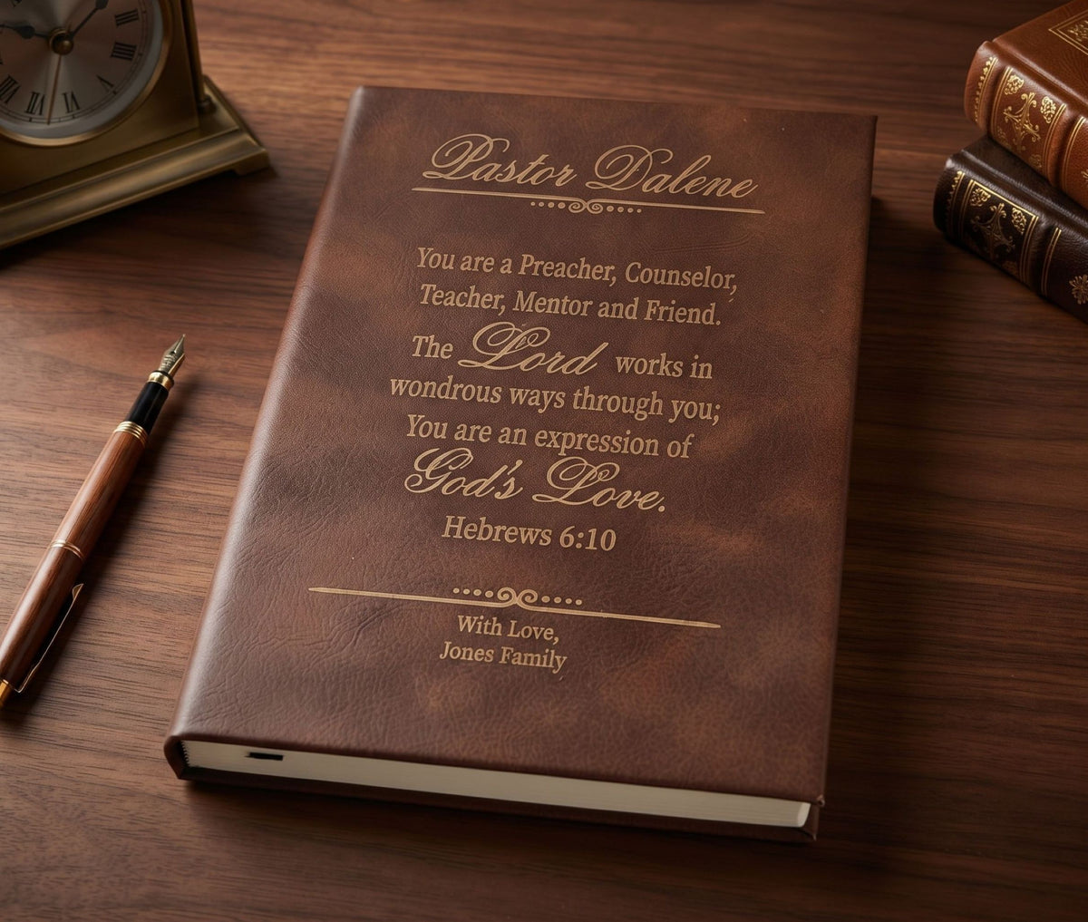 A personalized brown leather-bound book with gold-embossed text reading 'Pastor Daleáá' and a scripture quote, styled on a wooden desk with a pen, clock, and books.