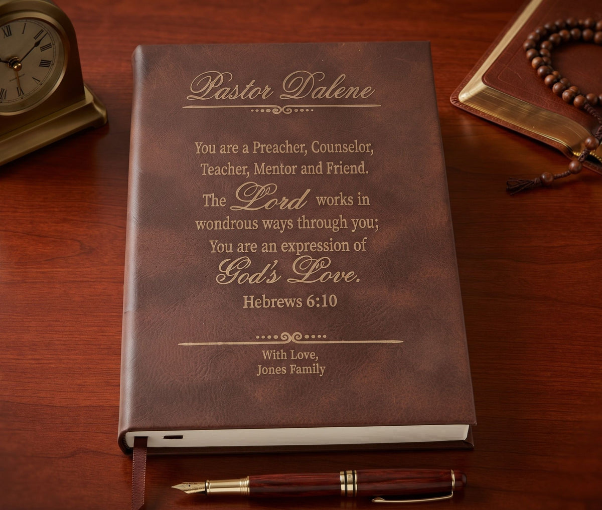 A personalized brown leather-bound Bible with gold foil lettering reading 'Pastor Dalene' and a scripture quote, displayed on a wooden desk next to a clock, pen, and rosary.