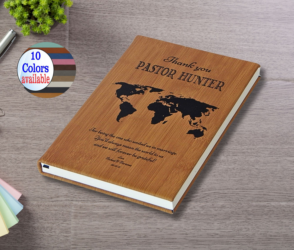 A personalized wooden keepsake journal with a laser-engraved world map and Bible verse, displayed on a gray wood-textured surface with a '10 Colors available' badge overlay.